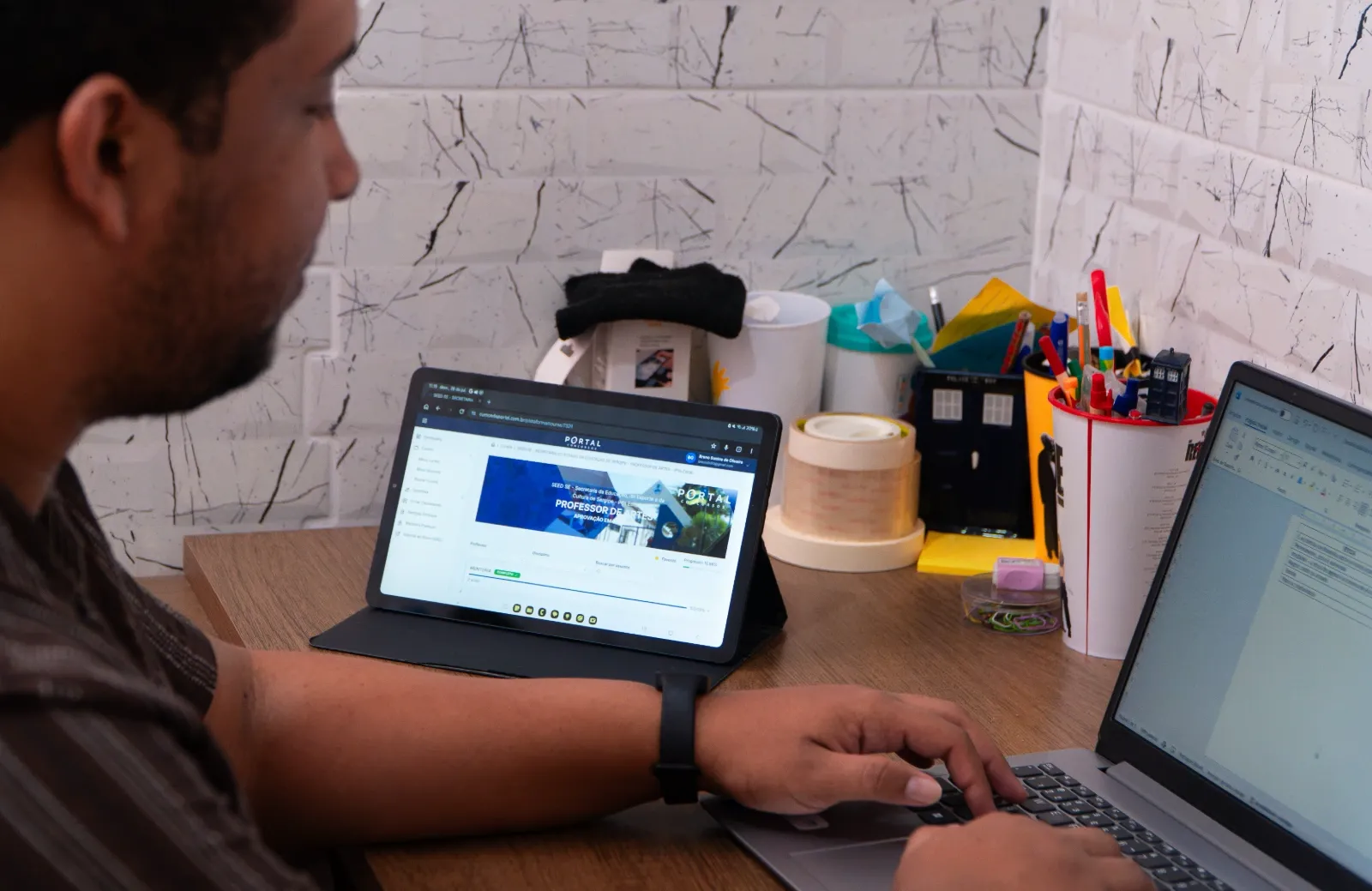 Pessoa do gênero masculino sentado diante de uma mesa de estudo com um tablet e um notebook logados na plataforma do Portal Concursos