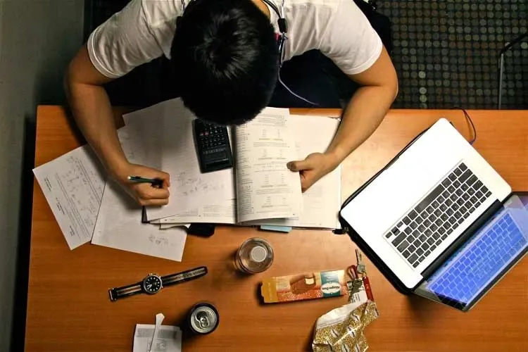 Garoto estudando à mesa com laptop, alguns itens pessoais, relógio, calculadora sobre o papel que segura e livros. Imagem dimensionada por uma posição de cima.